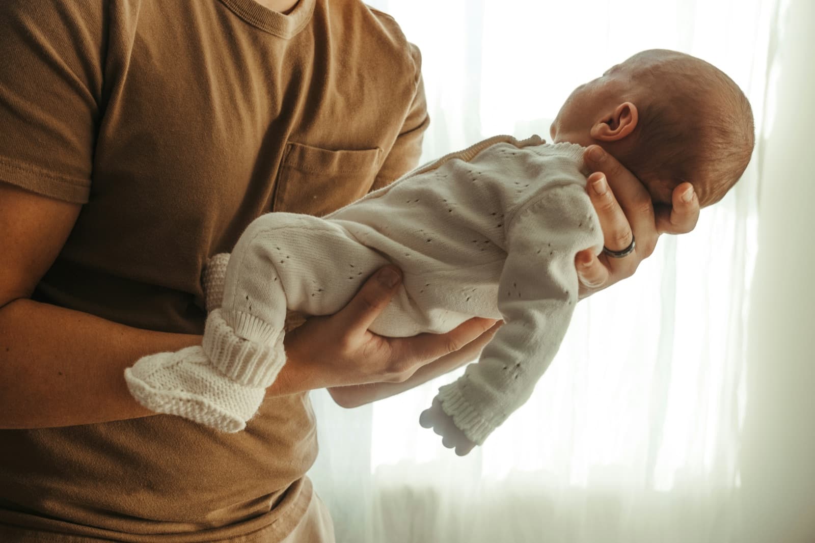 Mężczyzna w brązowej koszulce trzyma na przedramieniu niemowlę w białym, dzierganym pajacyku, w tle jasna firanka