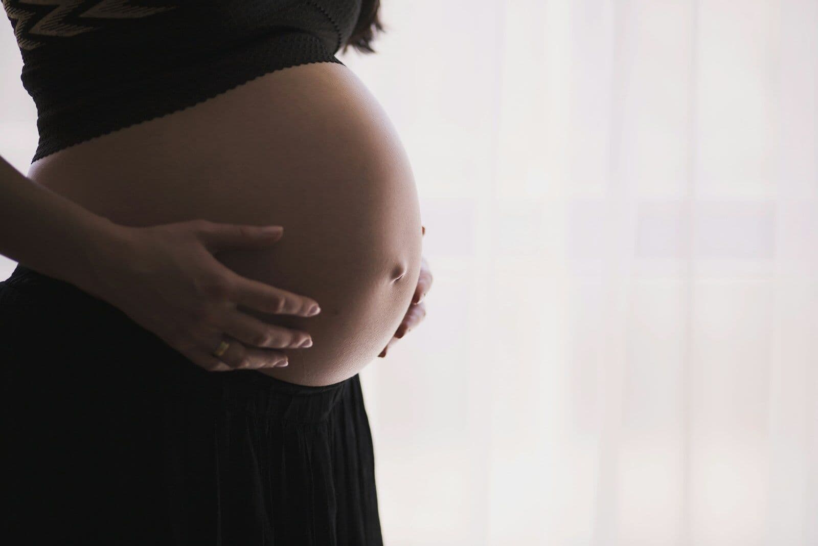 A heavily pregnant woman in a black skirt and a short black top stands side-on by a bright window with a net curtain, holding her bare belly in both hands
