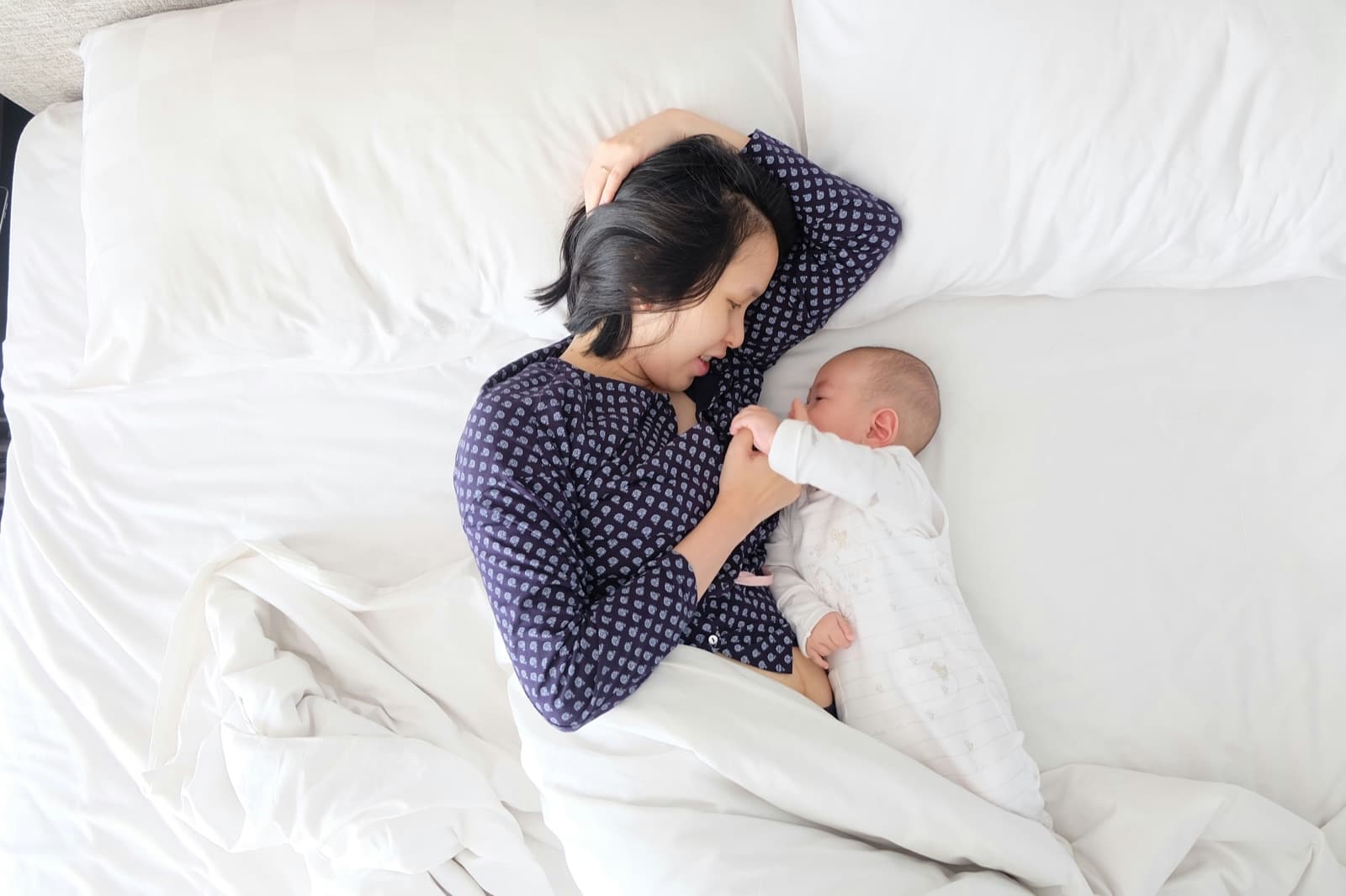 A woman in navy pyjamas lies on her side in white bedding, next to her lies a baby in a light sleepsuit whose hand she is holding