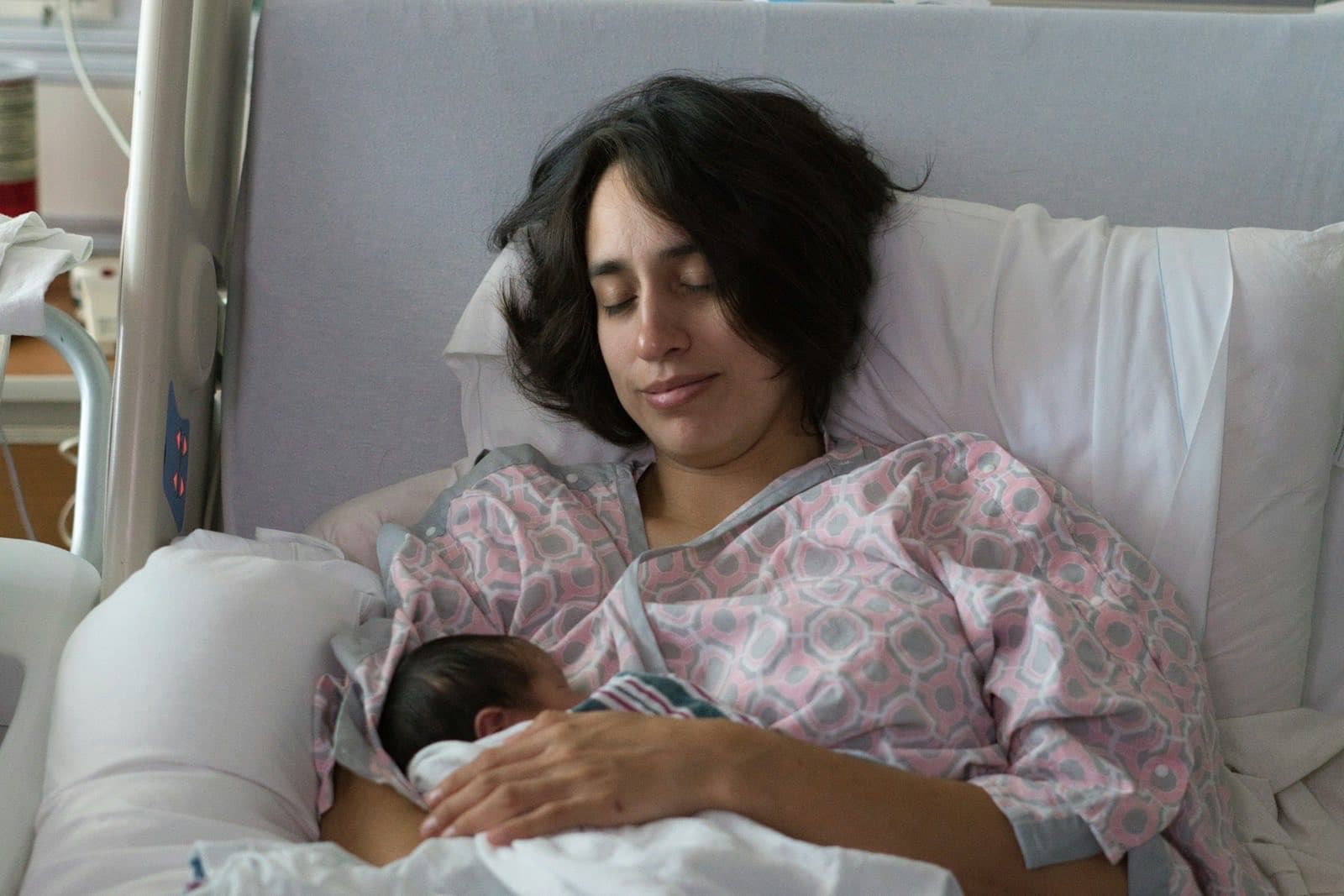 Mama w szpitalnej koszuli odpoczywa z zamkniętymi oczami na łóżku, na jej piersi śpi noworodek owinięty w pieluszkę