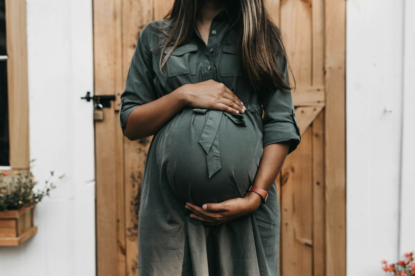 A heavily pregnant woman in a green shirt dress holds her belly with both hands in front of a wooden door