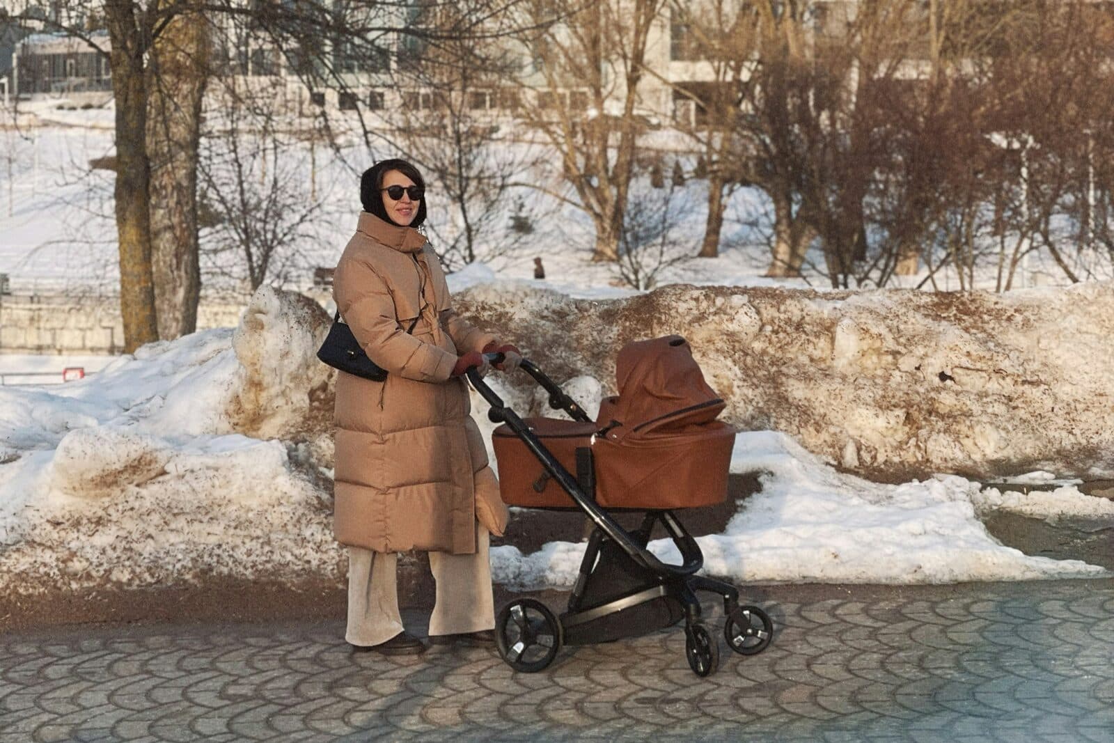 Жінка в довгому бежевому пуховику і темних окулярах стоїть із коричневим візком із глибокою люлькою на алеї в засніженому парку