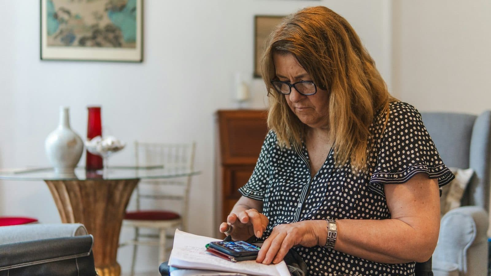 Kobieta w okularach i bluzce w groszki siedzi w fotelu w jasnym pokoju i liczy coś na telefonie, trzymając na kolanach plik zapisanych kartek